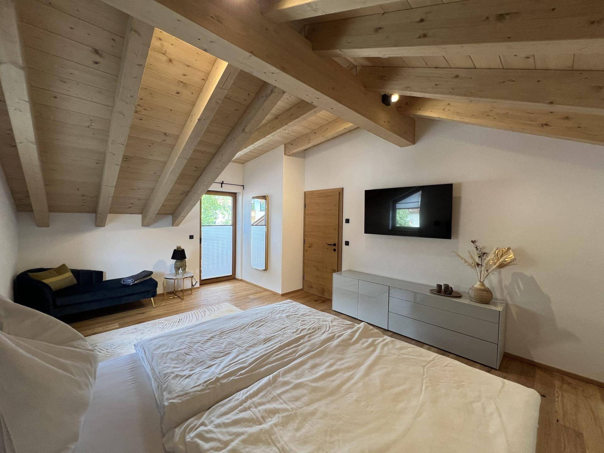 Helles Schlafzimmer mit Holzbalkendecke, Doppelbett, Kommode und Zugang nach draußen.