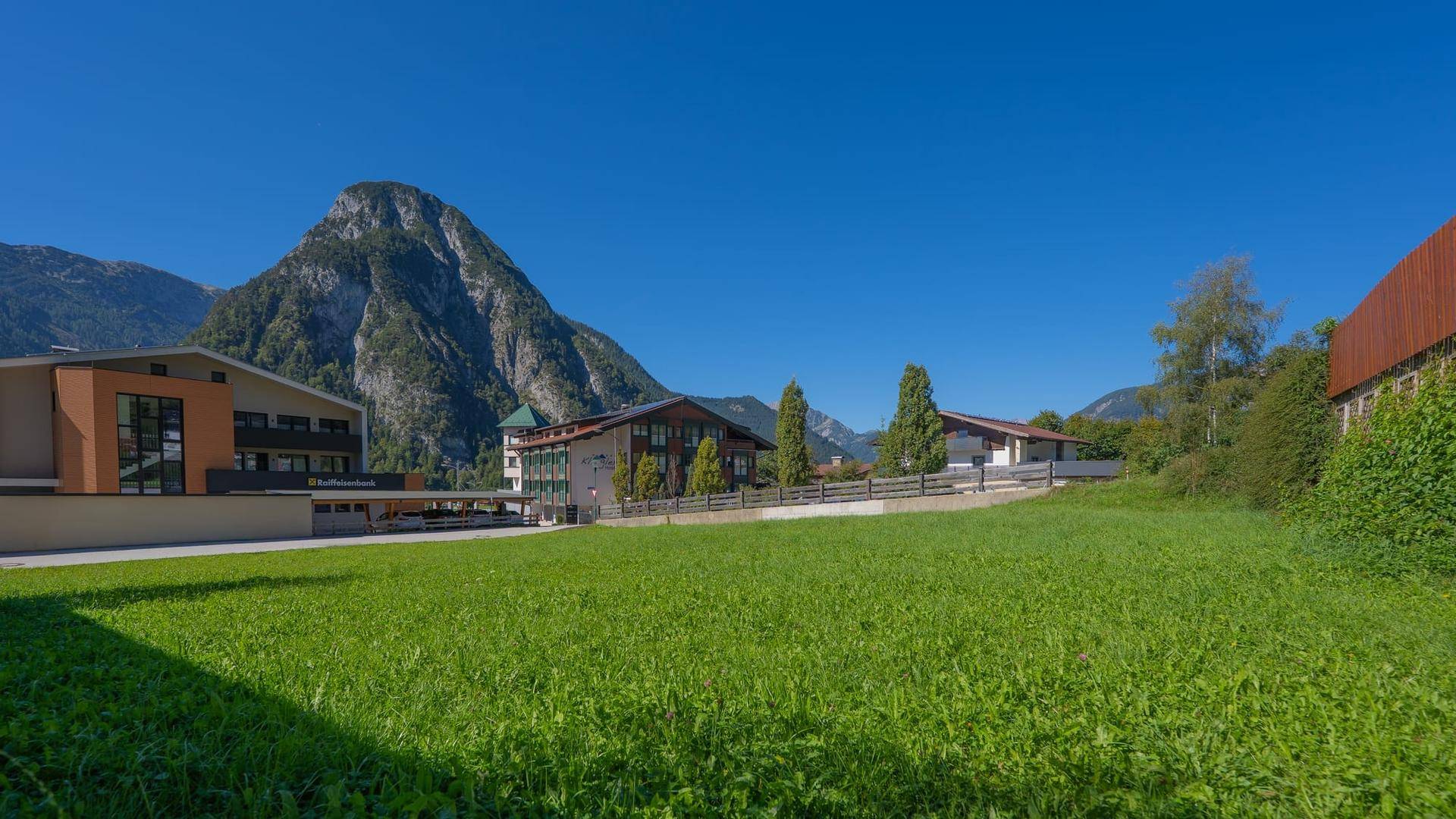 Panoramablick auf eine grüne Wiese mit Gebäuden und majestätischem Berg im Hintergrund unter blauem Himmel.