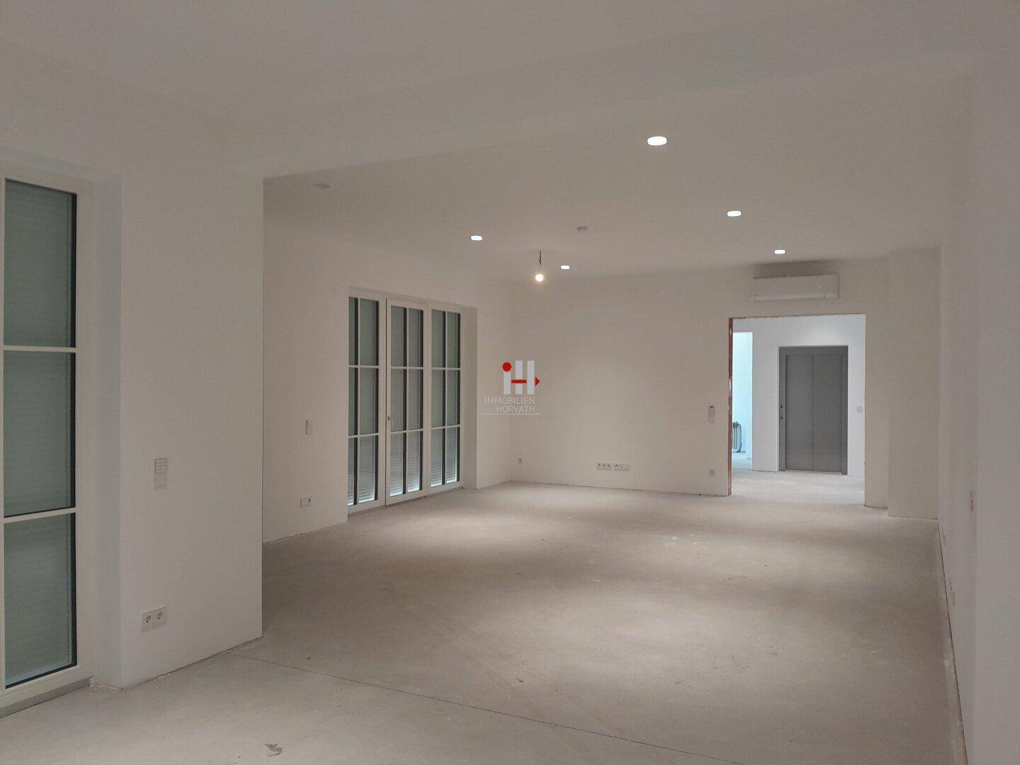 Offener Wohnbereich im Rohbau mit viel Tageslicht und vorbereiteter Klimaanlage.