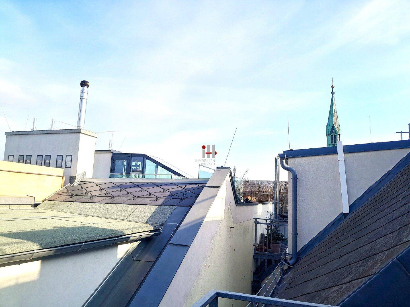 Dachlandschaft mit verschiedenen Gebäuden und einem Kirchturm unter blauem Himmel.
