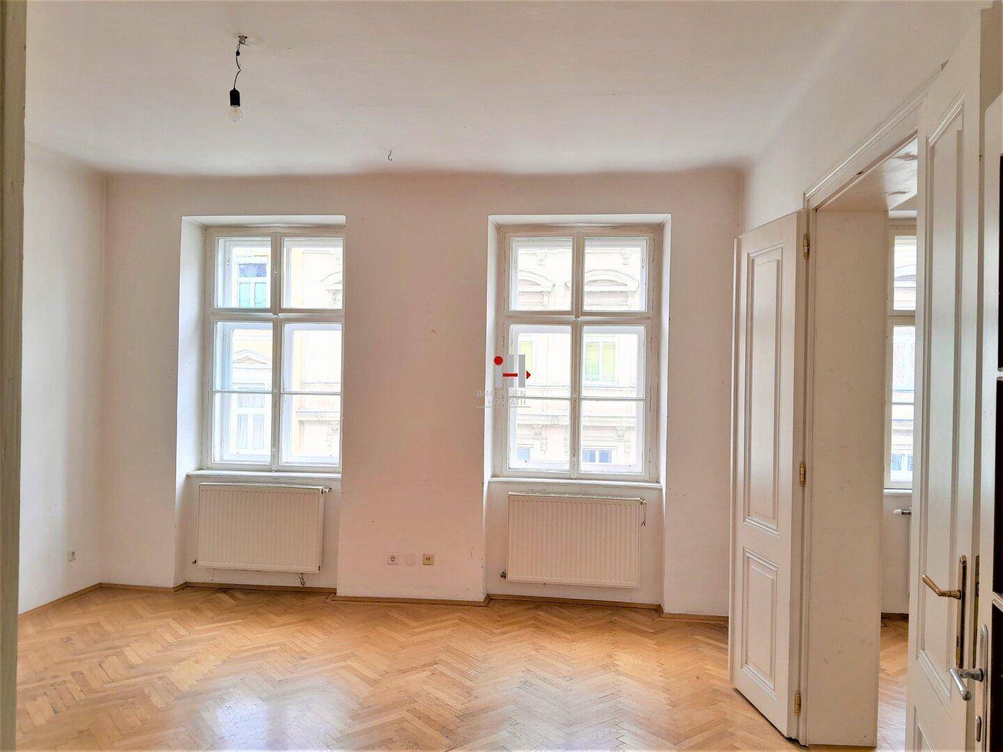 Geräumiger, heller Raum mit zwei großen Fenstern, Heizkörpern und klassischem Fischgrätparkett.