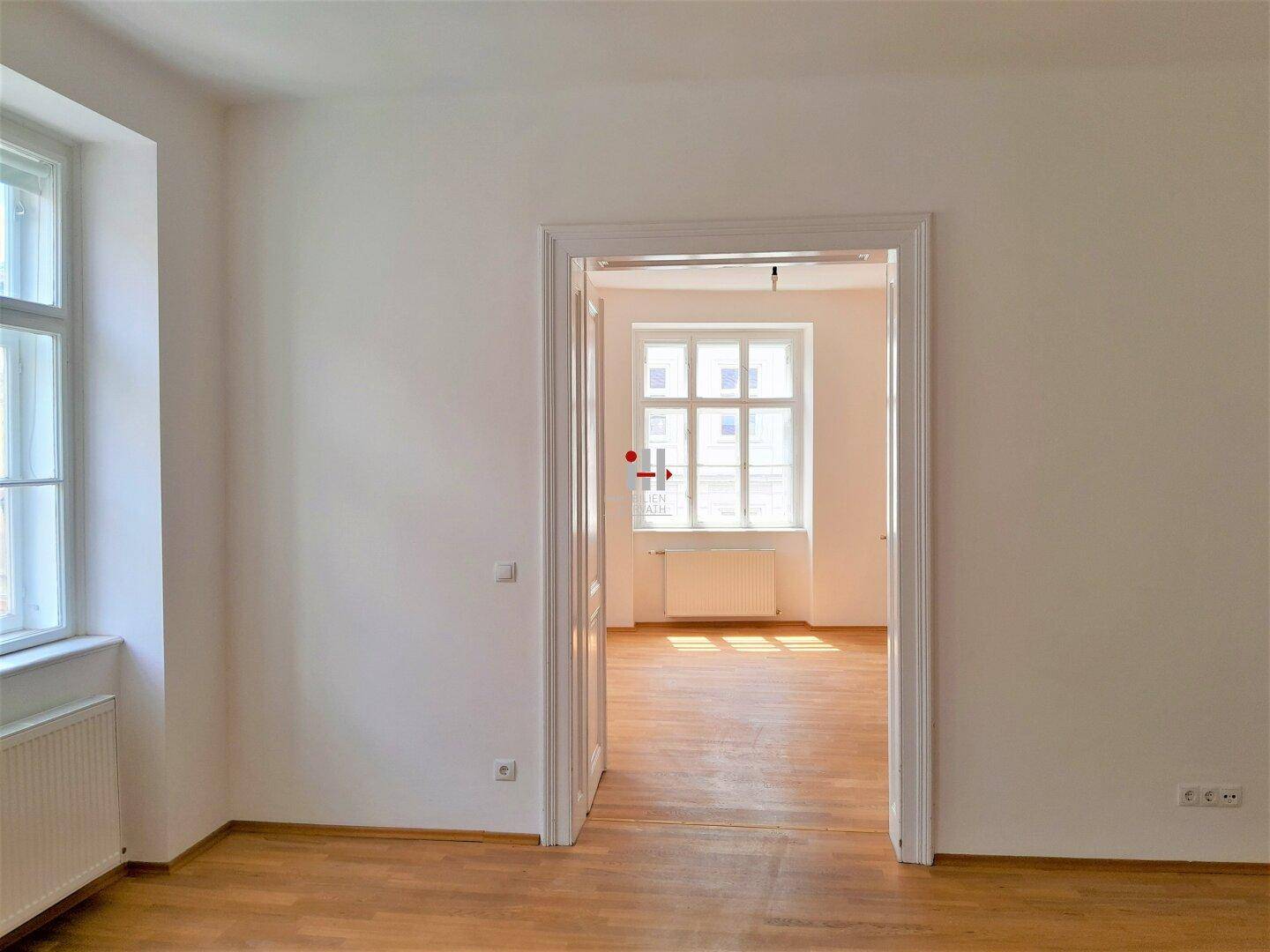 Geräumiger Raum mit hellem Fischgrätparkett und einem Fenster, das den Blick in ein weiteres Zimmer freigibt.