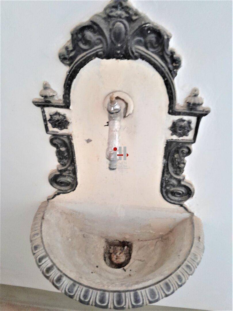 Historischer Wandbrunnen mit kunstvollen Verzierungen und einem Wasserhahn, möglicherweise in einem Flur.