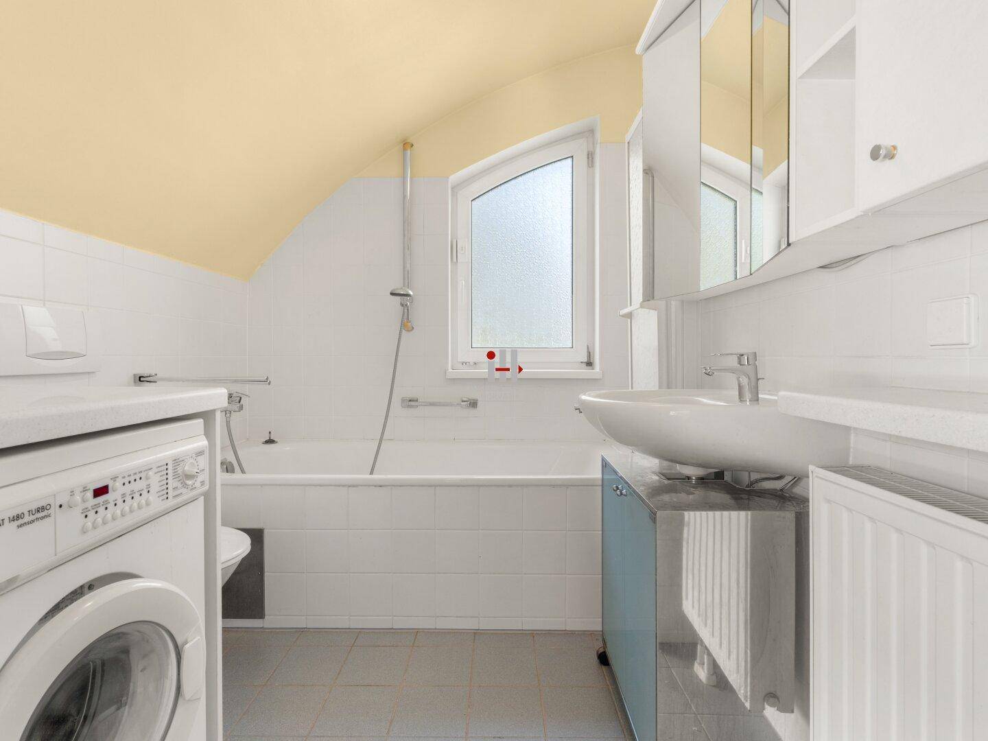 Badezimmer mit Badewanne, Waschbecken, Waschmaschine und Fenster unter Dachschrägen.