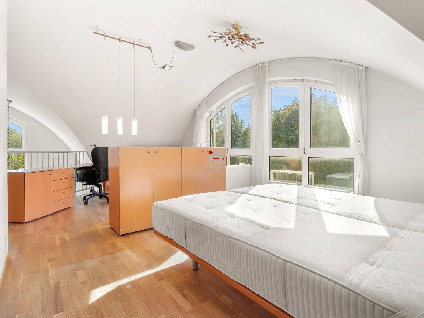 Helles Schlafzimmer mit Holzboden, großen Fenstern und Dachschrägen, offen zum Arbeitsbereich.