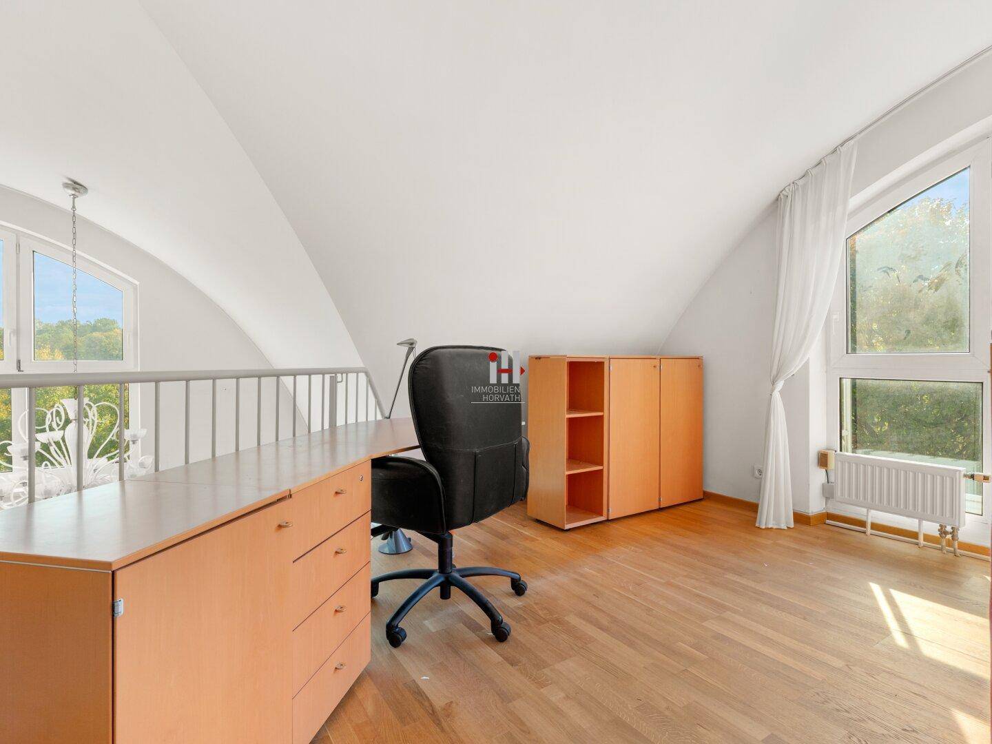 Heller Arbeitsbereich mit Holzboden, Dachschrägen und Blick ins Grüne durch große Fenster.