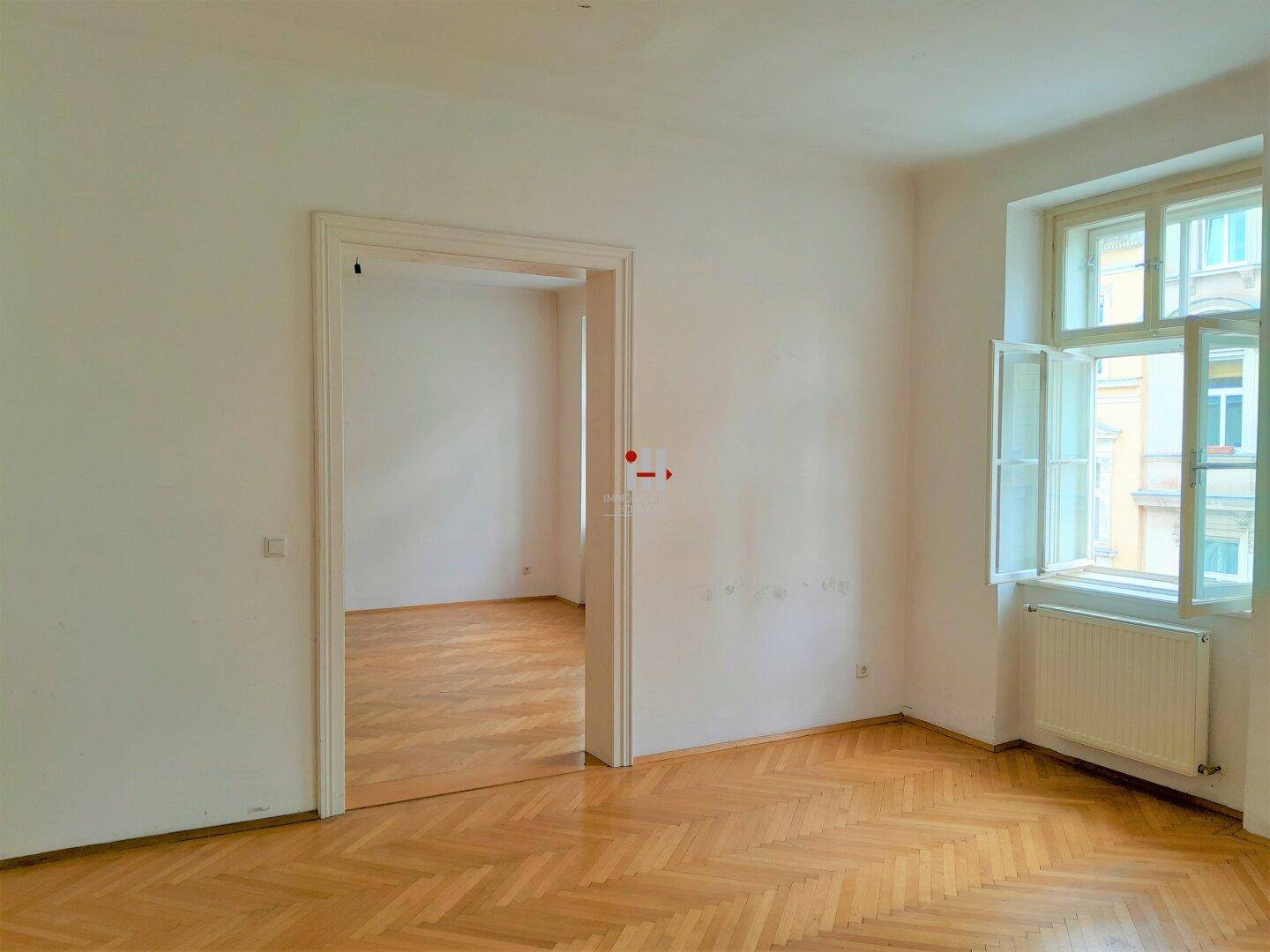 Leeres Zimmer mit Fischgrätparkett, weißen Wänden und einem großen Fenster mit Blick nach draußen.