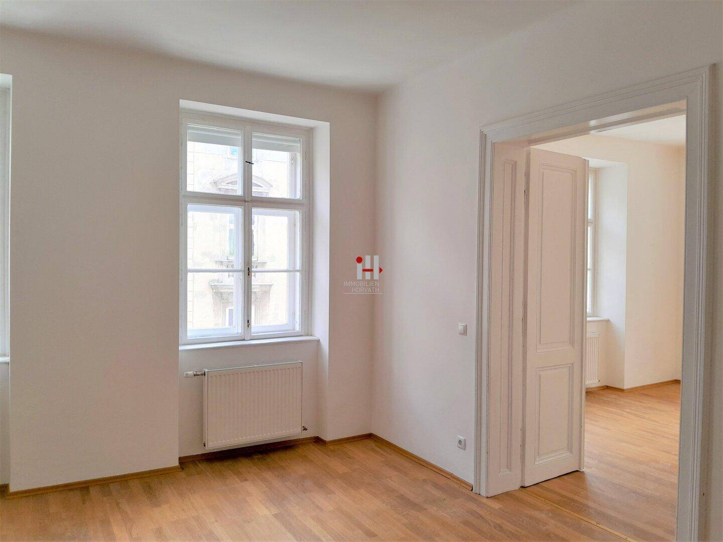 Helles, leeres Zimmer mit Parkettboden, hohen Fenstern und Flügeltüren zu einem angrenzenden Raum.