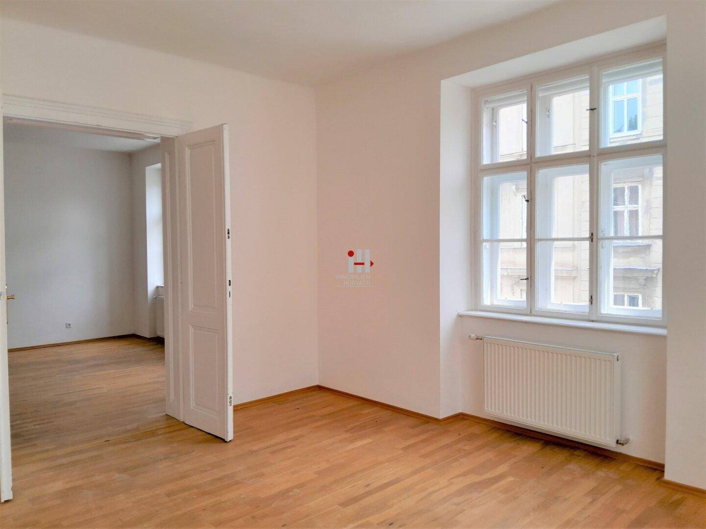 Lichtdurchflutetes Zimmer mit Parkettboden, weißen Wänden und einem Heizkörper unter dem Fenster.