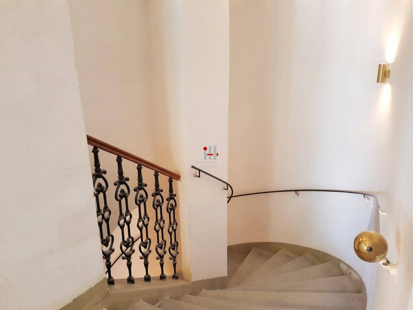Historisches Treppenhaus mit geschwungenen Stufen, schmiedeeisernem Geländer und Wandbeleuchtung.