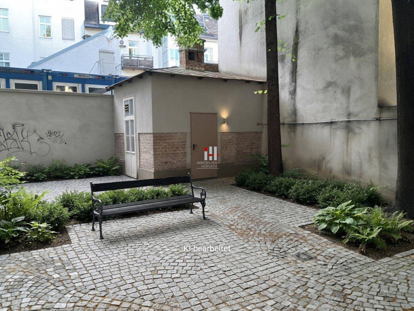 Gepflasterter Innenhof mit Sitzbank, umgeben von Grünflächen und einem kleinen Nebengebäude.