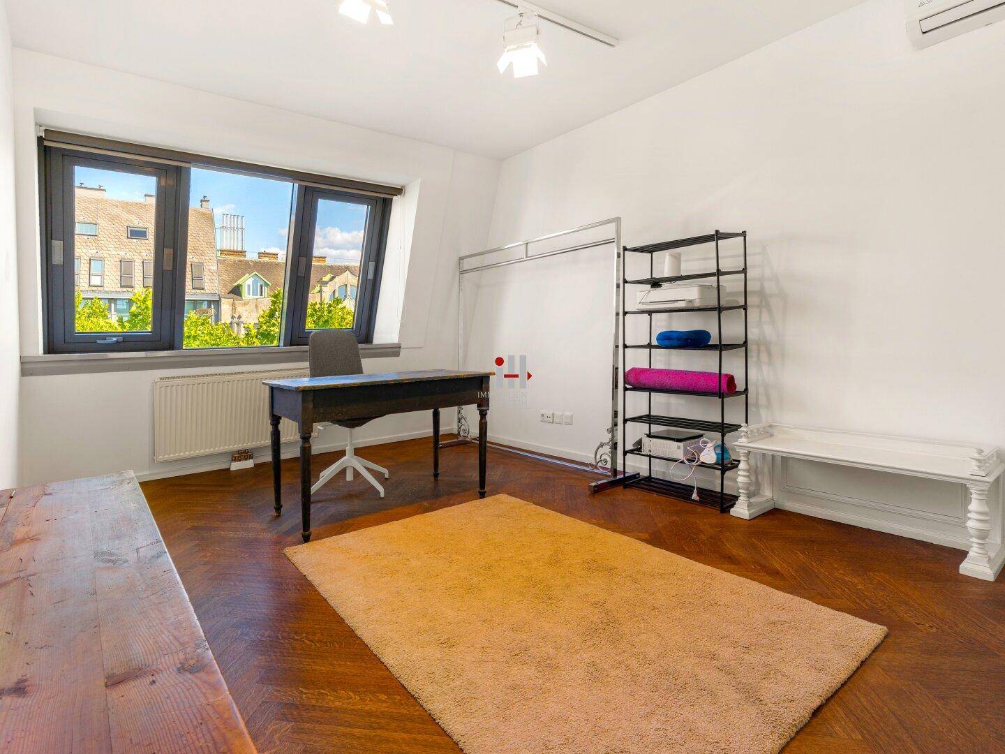 Helles Arbeitszimmer mit Parkettboden, Schreibtisch und Blick auf die umliegenden Dächer.