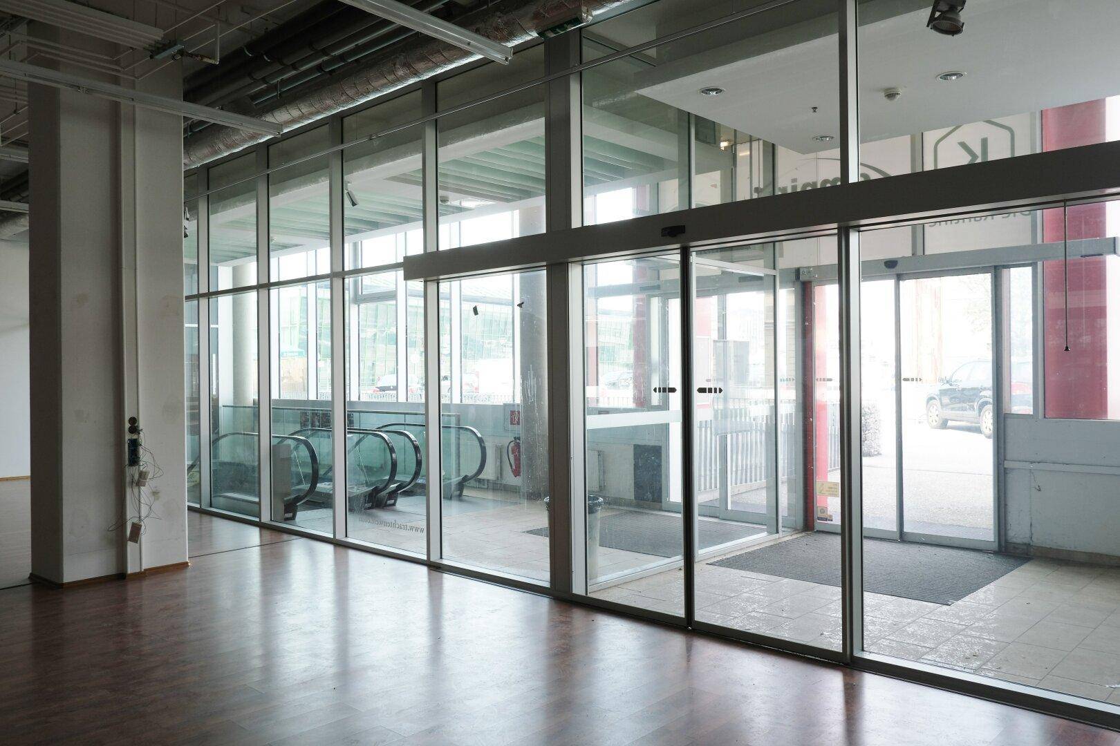 Heller Eingangsbereich mit großen Glasfronten, automatischen Türen und Rolltreppen im Hintergrund.