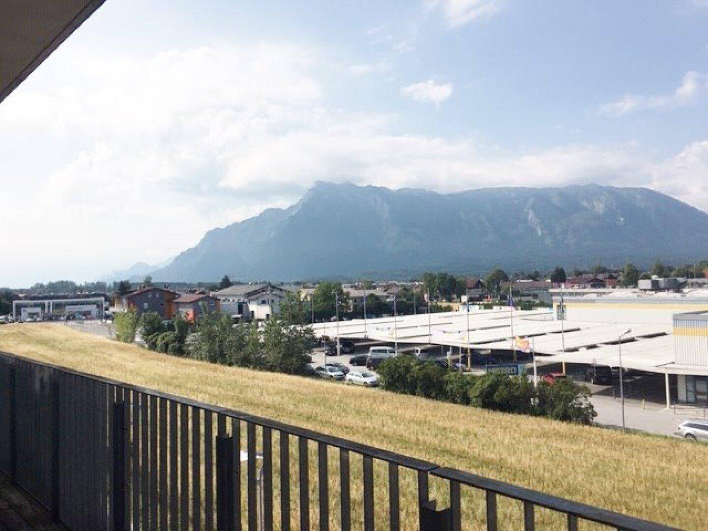 Weitblick von einem Balkon über eine Gewerbefläche mit Parkplätzen und einer beeindruckenden Bergkulisse im Hintergrund.