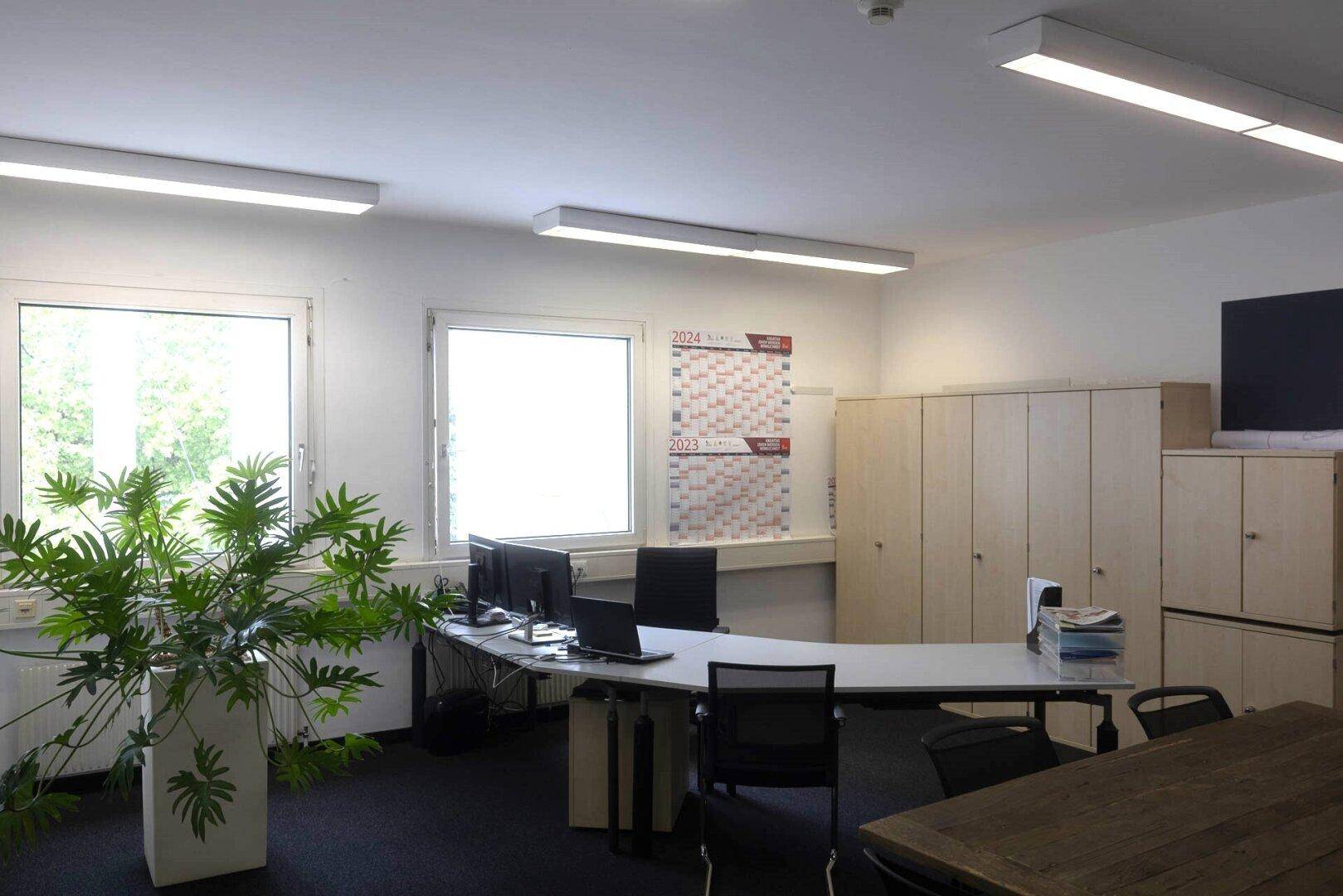 Helles Büro mit zwei Fenstern, Schreibtischen, Computern und hellen Holzschränken.