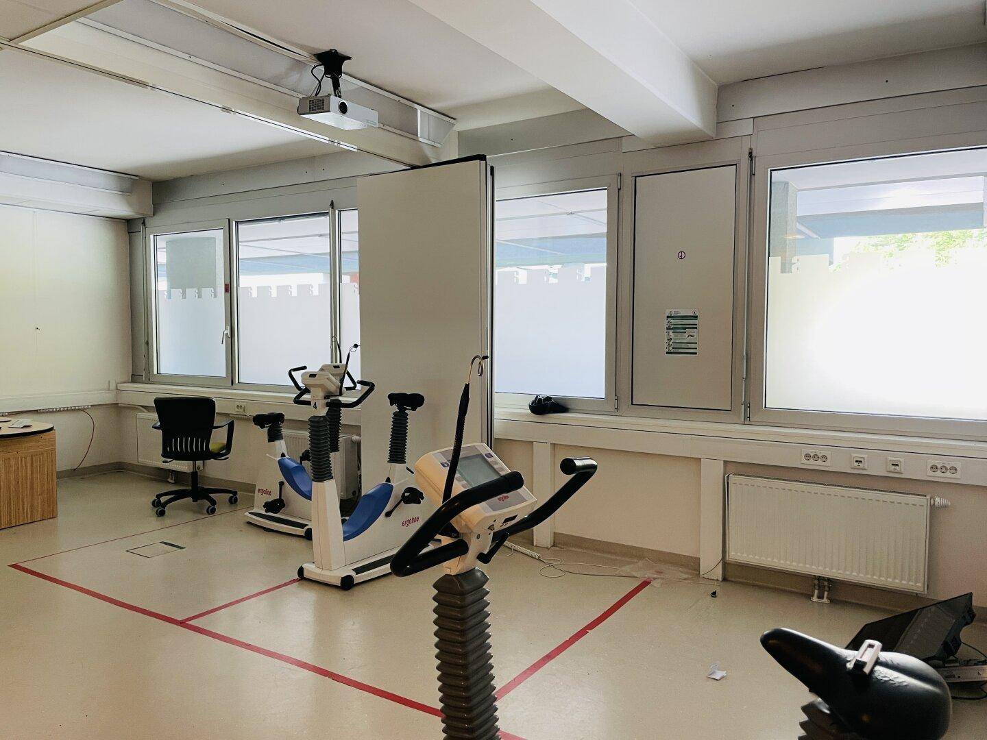 Trainingsraum mit mehreren Ergometern, großen Fenstern und einem Projektor an der Decke.