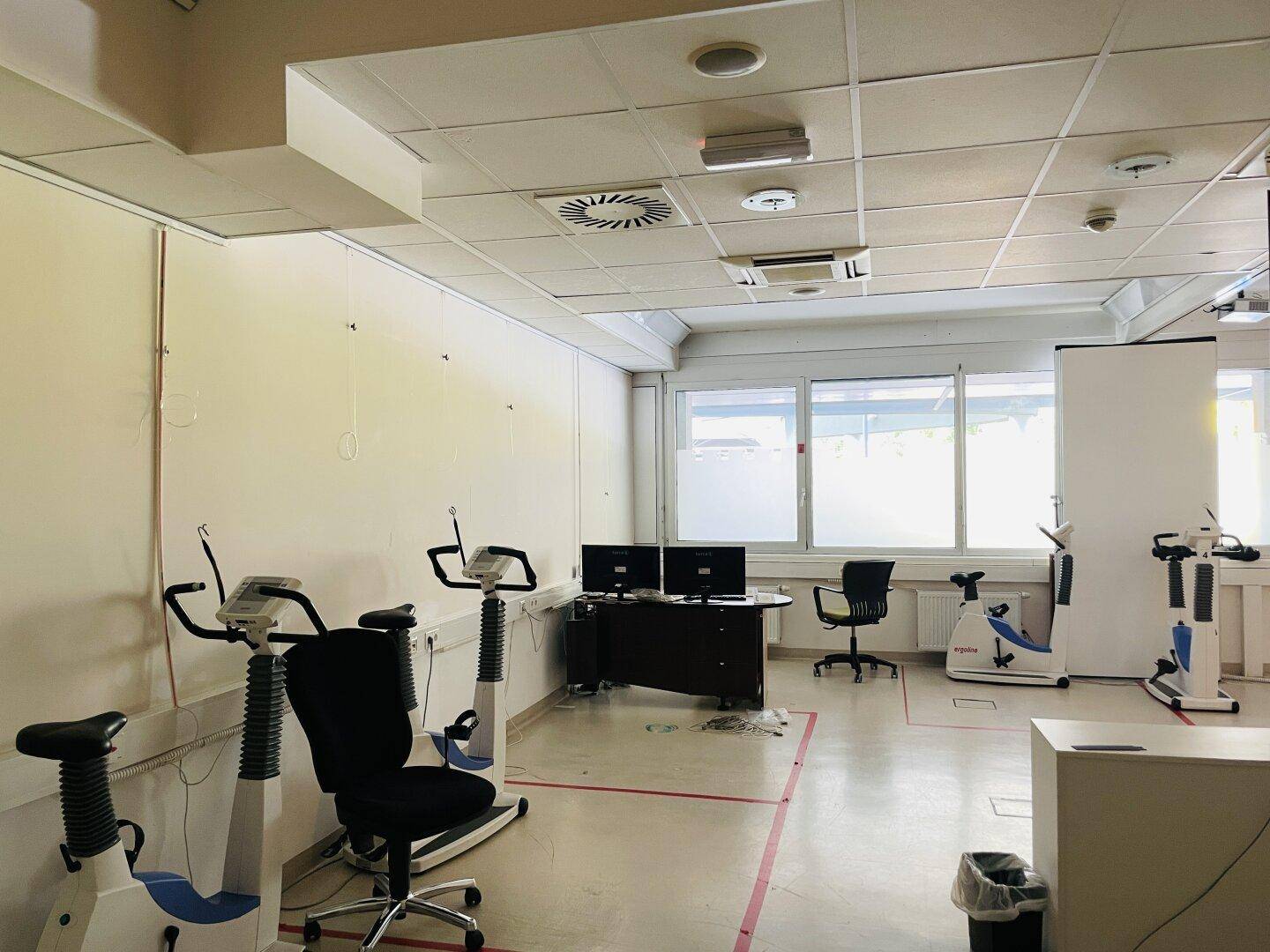 Trainingsraum mit verschiedenen Fitnessgeräten, Monitoren und großen Fenstern für Tageslicht.
