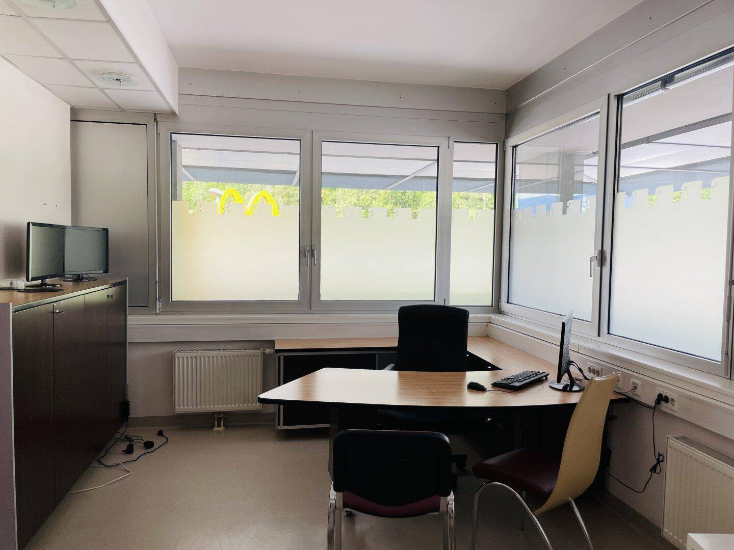 Büroraum mit Schreibtisch, Bürostuhl, Computermonitoren und großen Fenstern mit Sichtschutz.