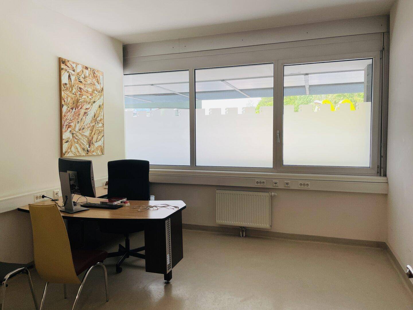 Büro mit Schreibtisch, Stühlen und Computermonitoren, beleuchtet durch große Fenster.