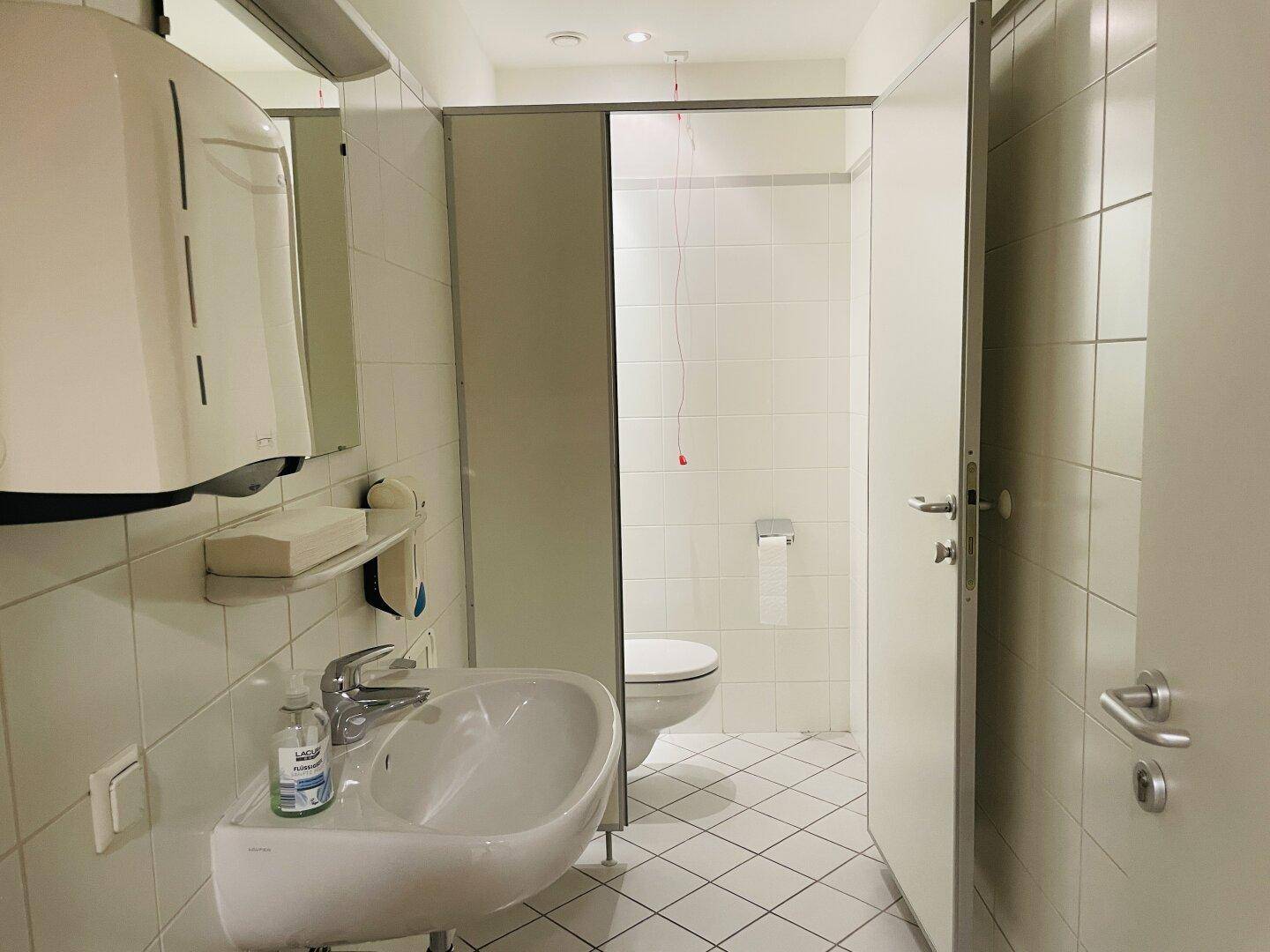 Funktionelles Badezimmer mit Waschbecken, Toilettenkabinen und weißen Fliesen an Wänden und Boden.