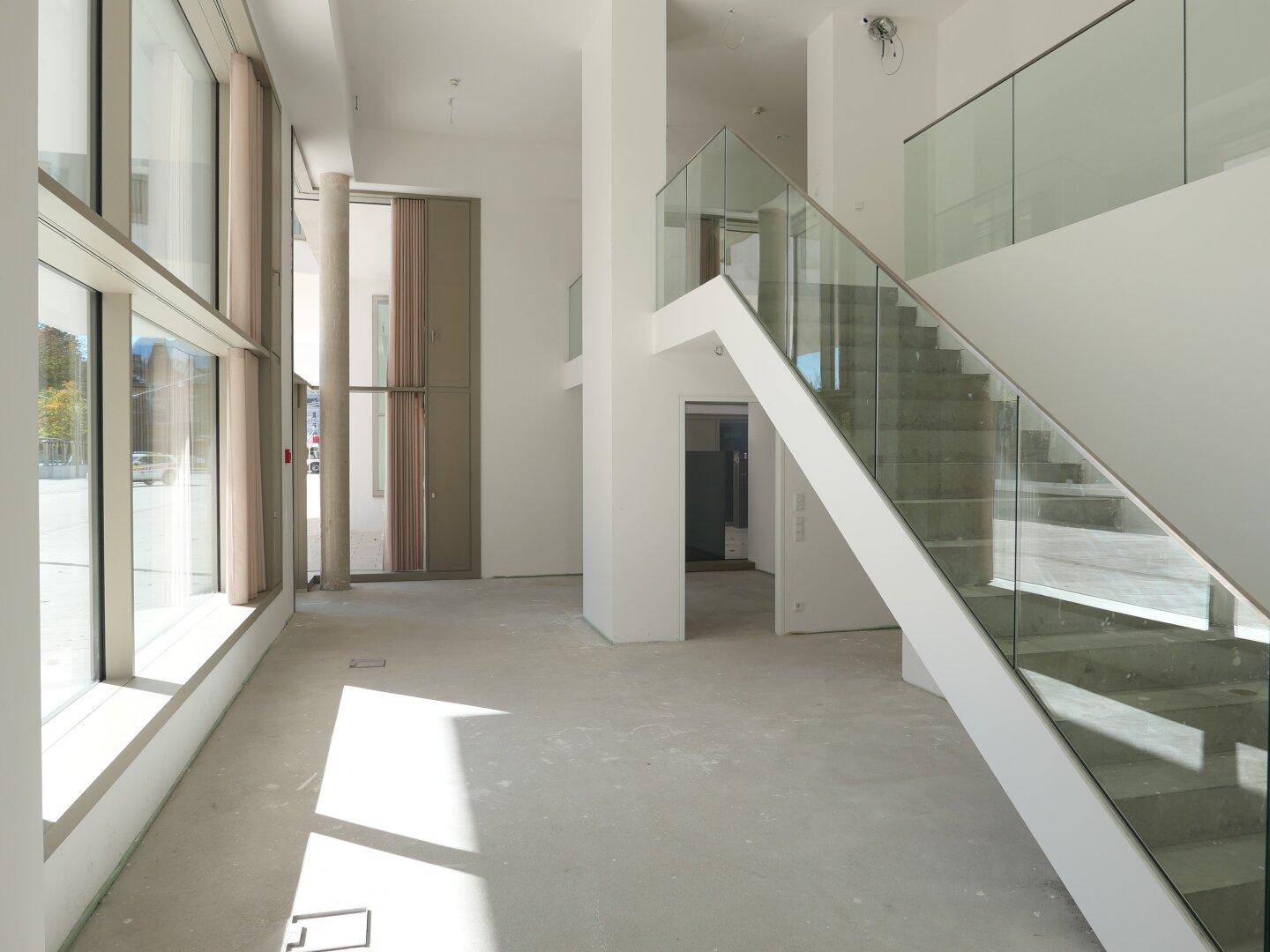 Geräumiger Eingangsbereich mit moderner Glasgeländer-Treppe und viel Tageslicht durch große Fenster.