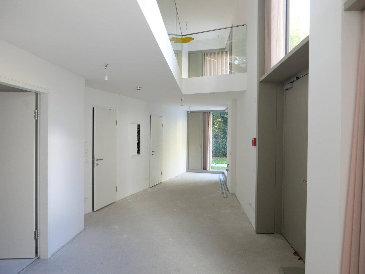 Langer, heller Flur in einem modernen Gebäude mit Aufzug und Blick ins Grüne durch große Fenster.
