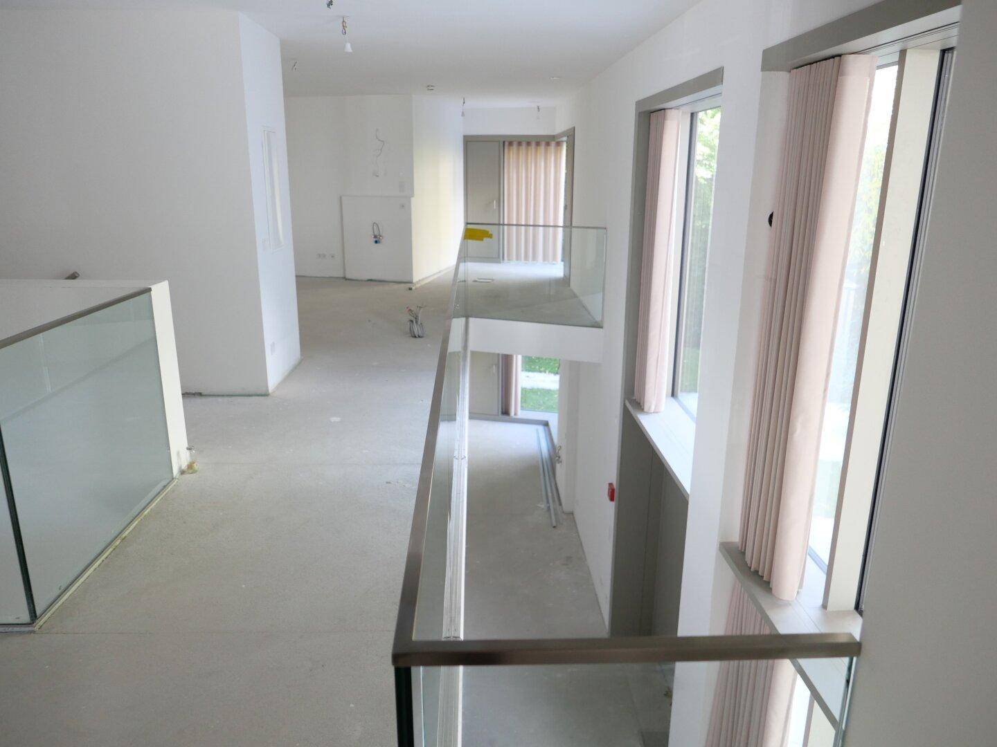 Heller Flur in einem Neubau mit Glasgeländer, hohen Decken und großen Fenstern für viel Tageslicht.