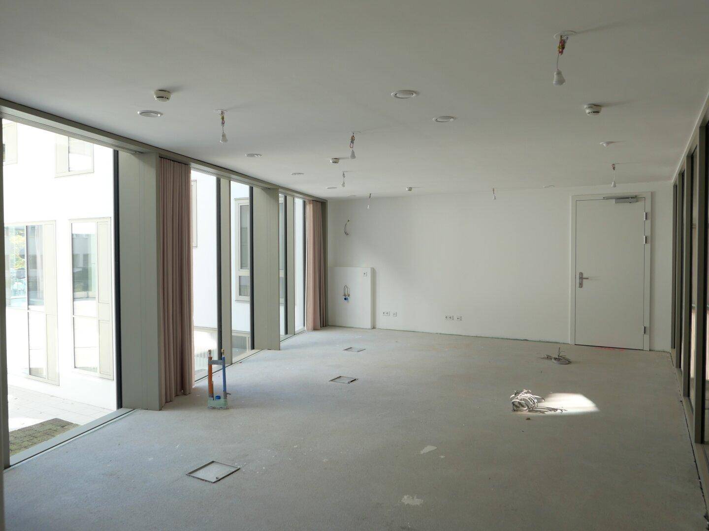 Großer, leerer Raum in einem Neubau mit bodentiefen Fenstern, viel Tageslicht und Betonboden.