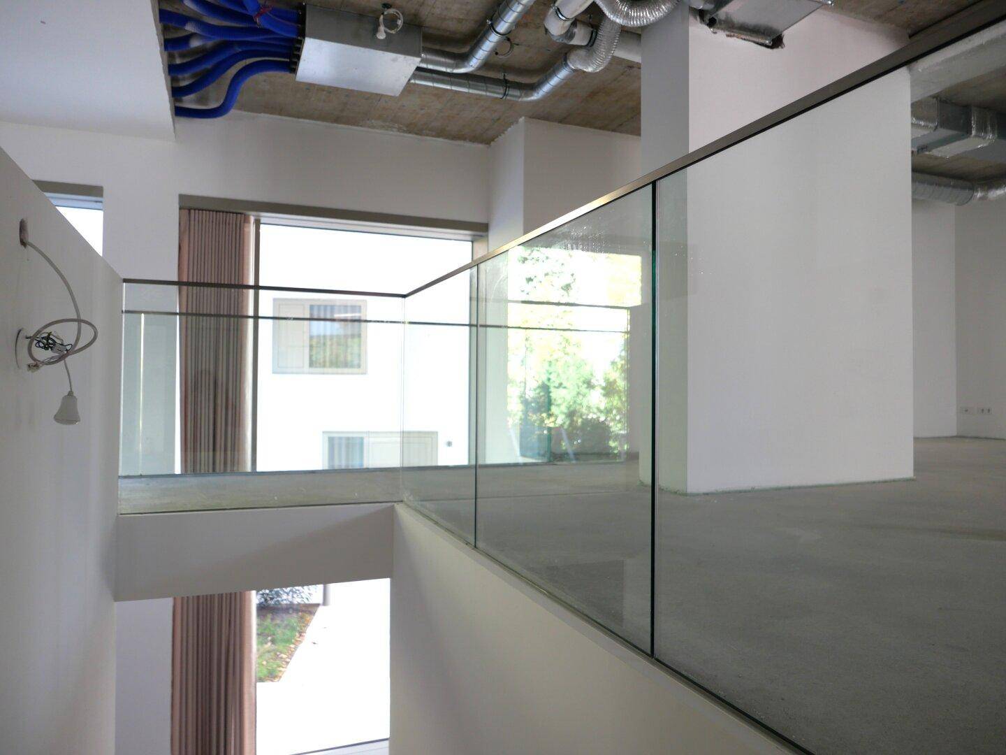 Offener Innenbereich mit Glasgeländer, hohen Decken und sichtbaren Lüftungsrohren im Industriedesign.