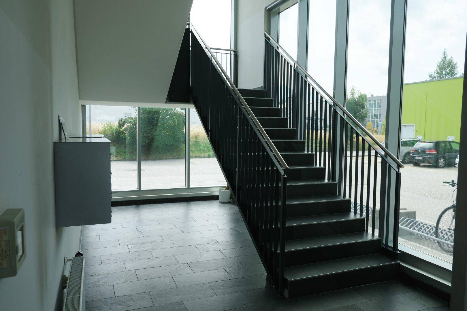 Modernes Treppenhaus mit dunklen Stufen, Metallgeländer und großen Fenstern mit Blick nach draußen.
