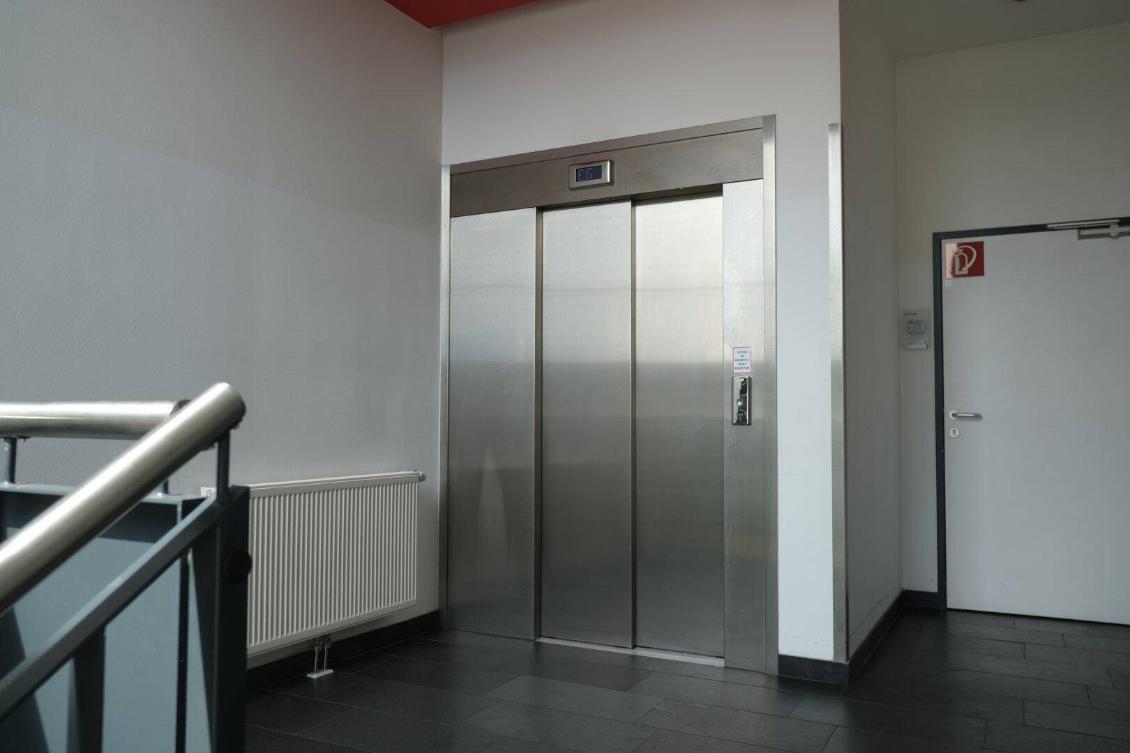 Moderner Aufzug mit Edelstahltüren in einem Flur mit dunklem Fliesenboden und Heizkörper.
