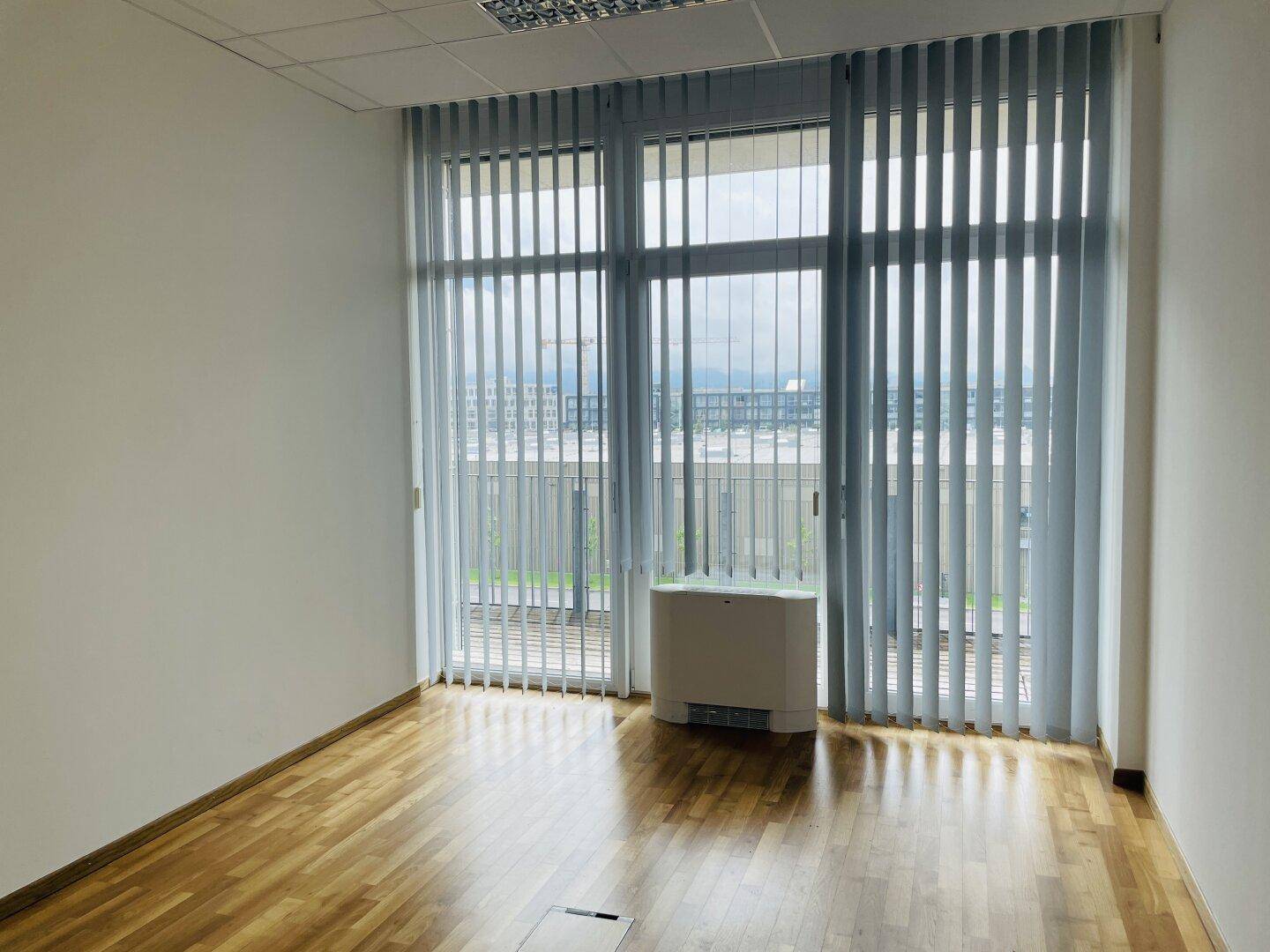 Heller Büroraum mit Holzboden, weißen Wänden und vertikalen Jalousien an den großen Fenstern.