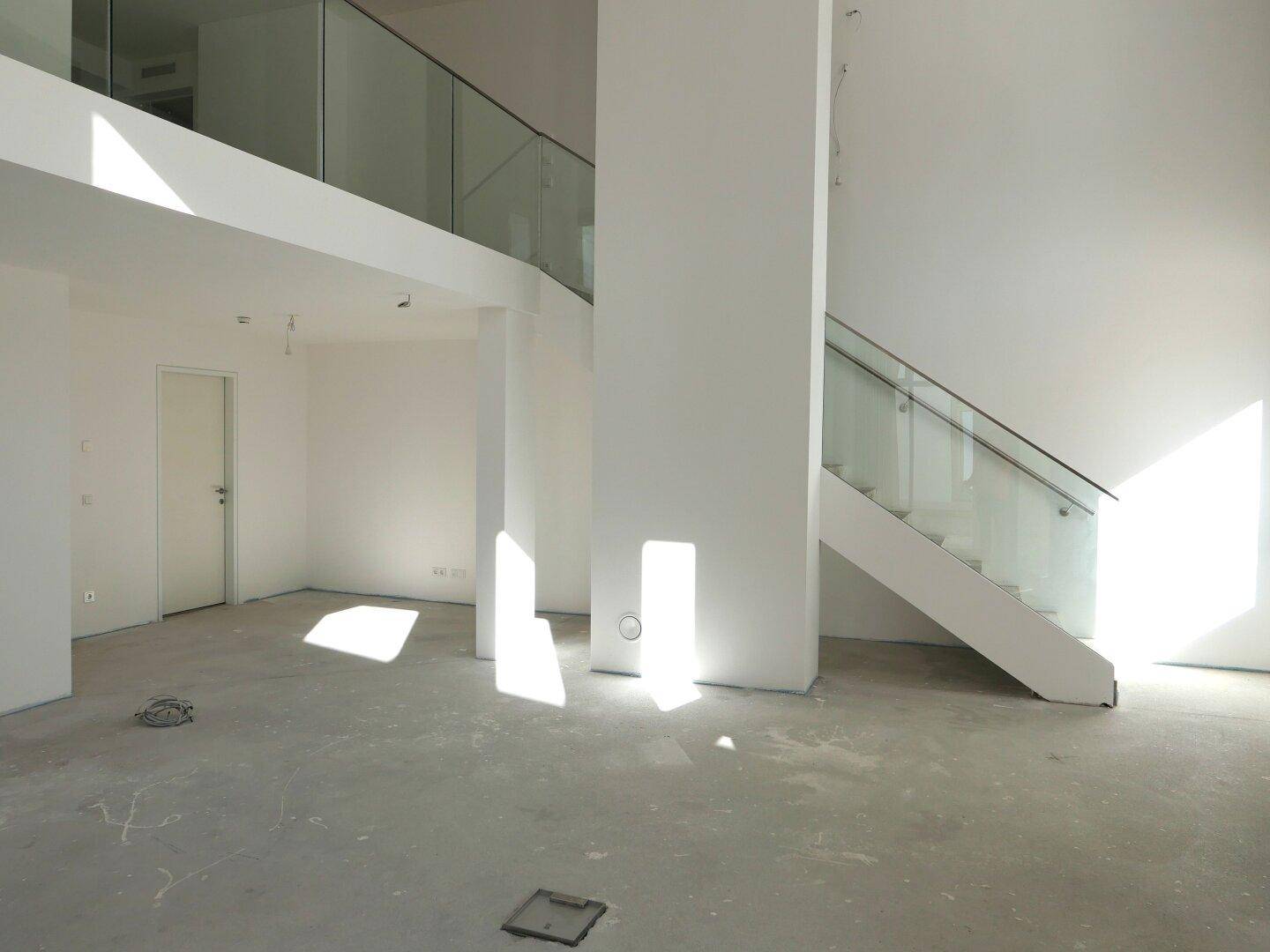 Heller, unfertiger Innenraum mit Betondecke, Treppe mit Glasgeländer und viel Tageslicht.