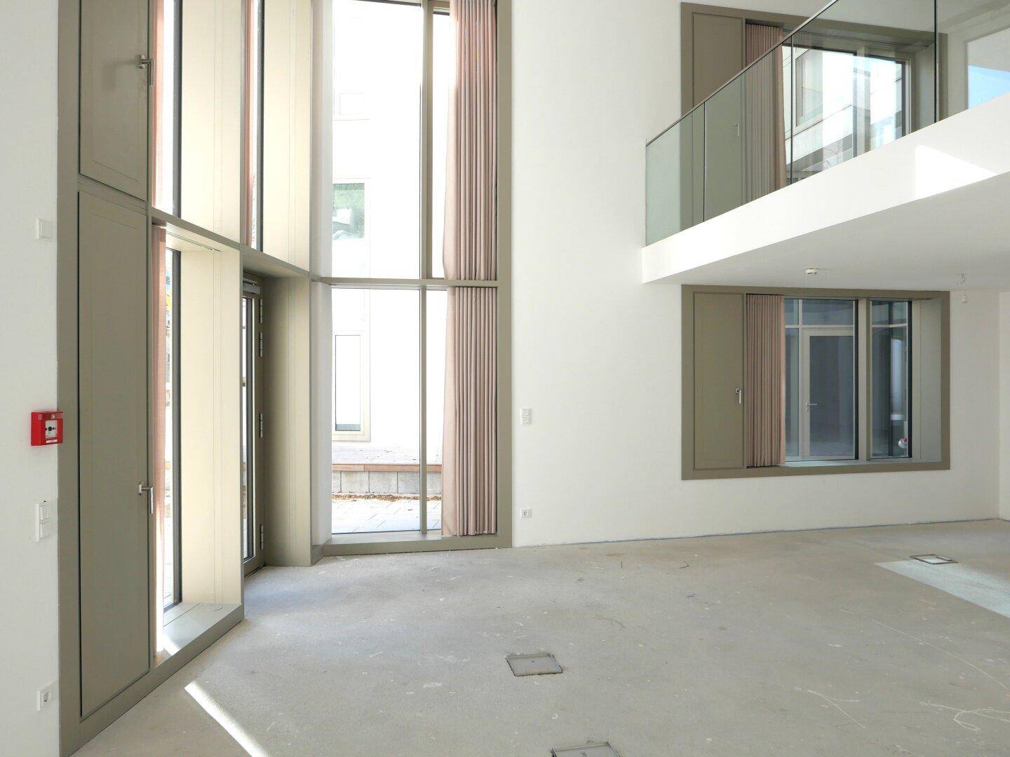 Unfertiger Innenraum mit hohen Fenstern, einer Galerie und einem roten Feuerlöscher an der Wand.