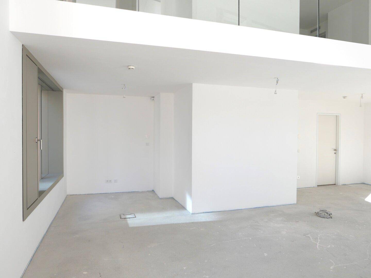 Leerer, heller Raum mit Betondecke, einem Fenster und einer Galerieebene im Hintergrund.