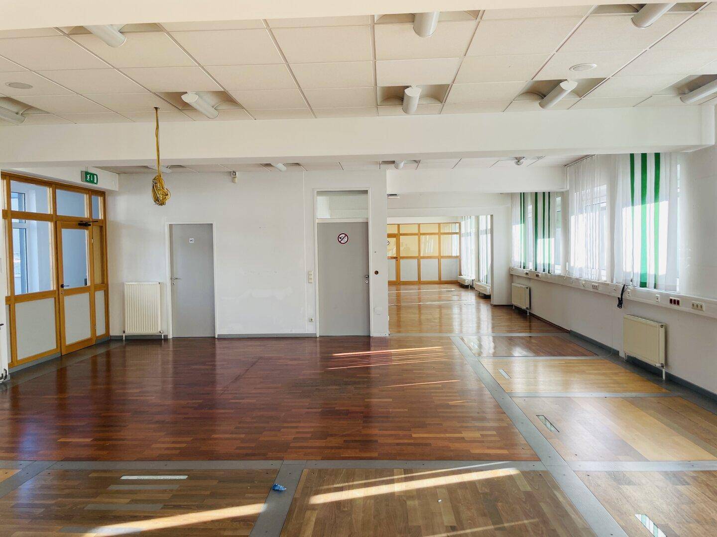 Großer, leerer Raum mit Holzparkettboden, vielen Türen und Fenstern mit vertikalen Jalousien, bereit zur Gestaltung.