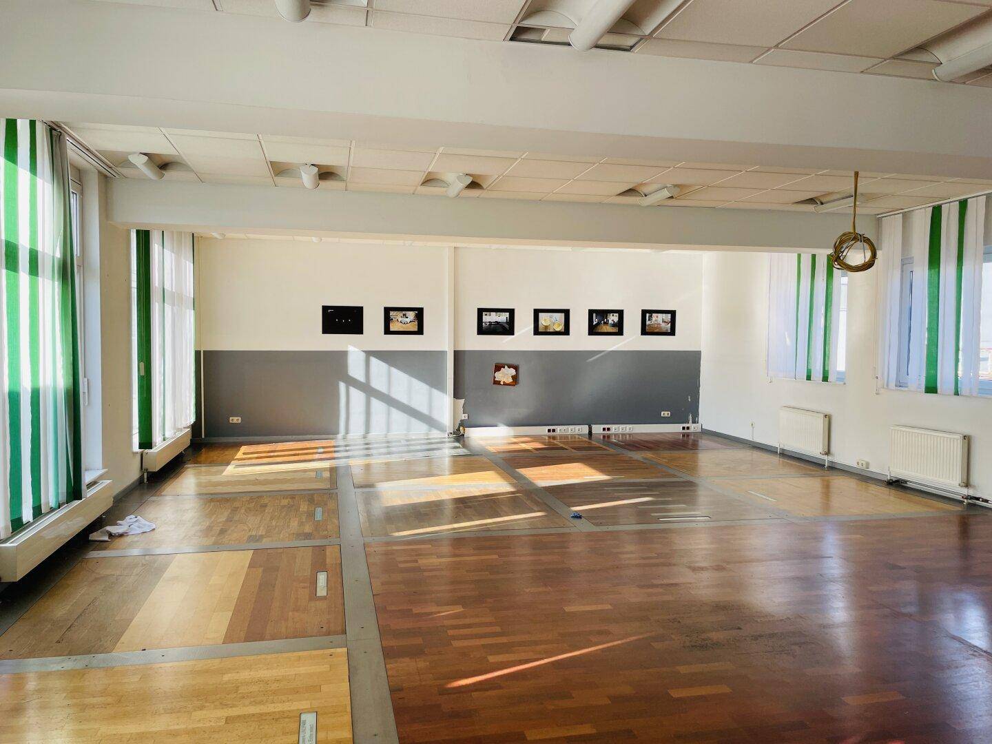 Heller, weitläufiger Raum mit Holzparkettboden, hohen Decken und Fenstern mit grünen Jalousien und Kunst an der Wand.