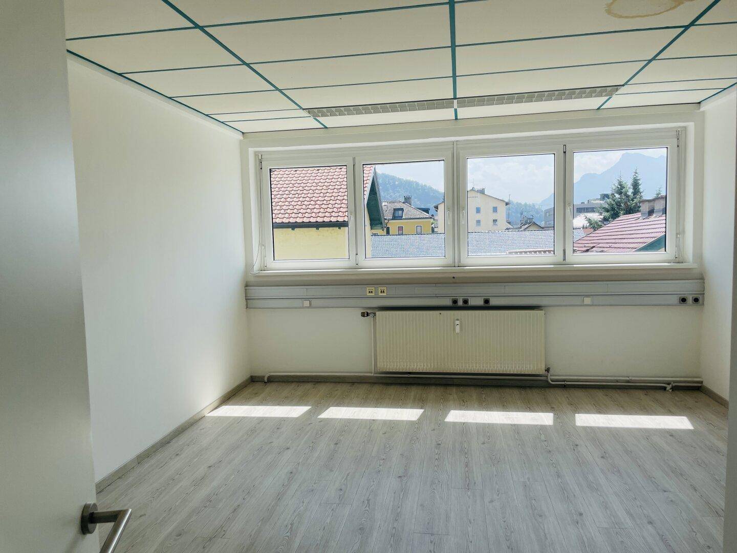 Geräumiges Büro mit hellem Laminatboden, Fensterfront und Ausblick auf die umliegende Landschaft und Berge.