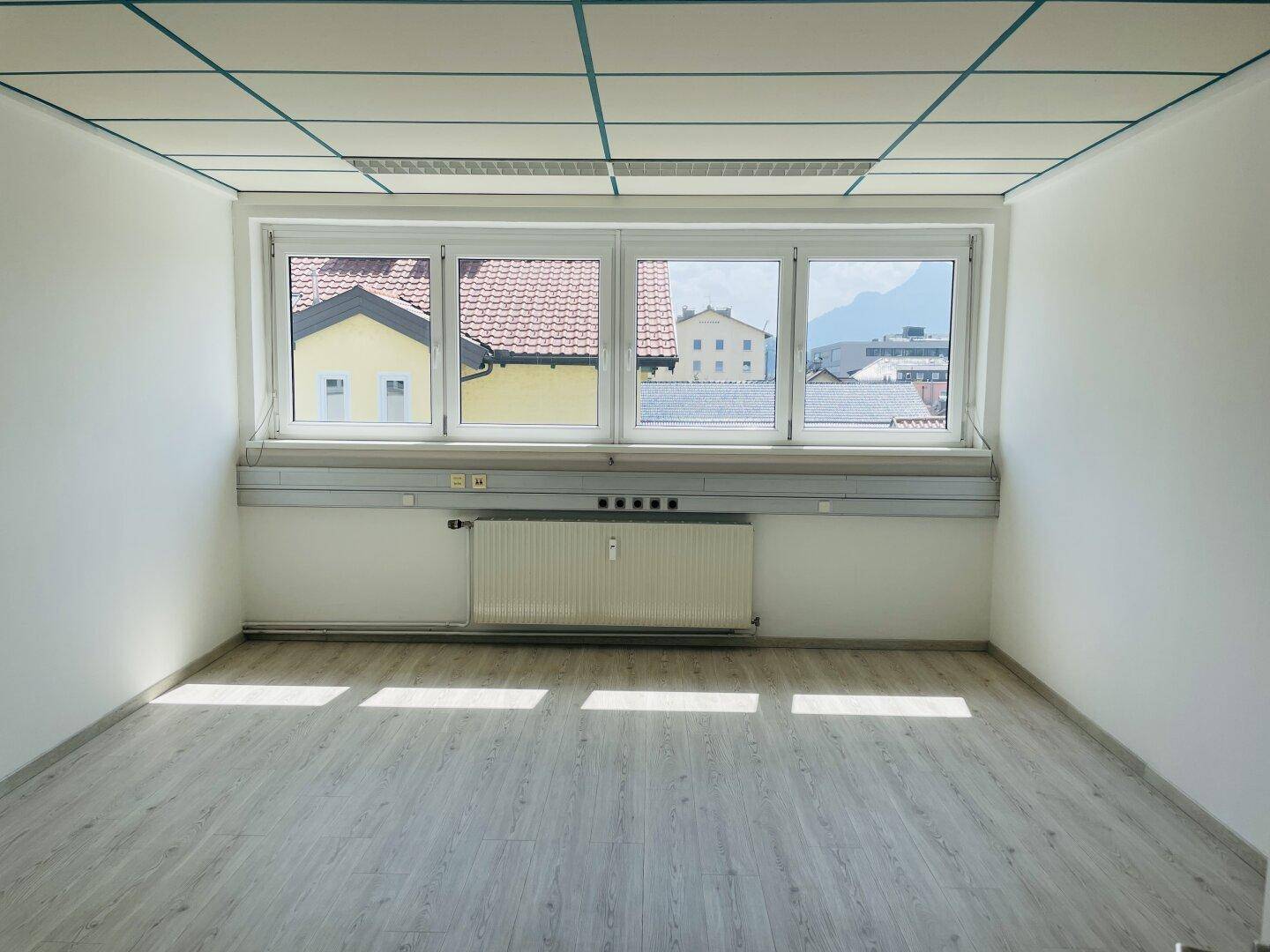 Helles Büro mit Laminatboden, großem Fenster und Blick auf Häuser, grüne Landschaft und Berge.