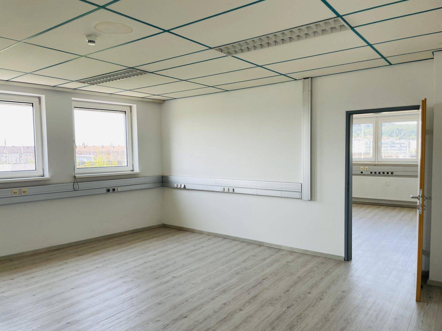 Heller Büroraum mit Laminatboden und Rasterdecke, mit Zugang zu einem weiteren Bereich durch eine offene Tür.