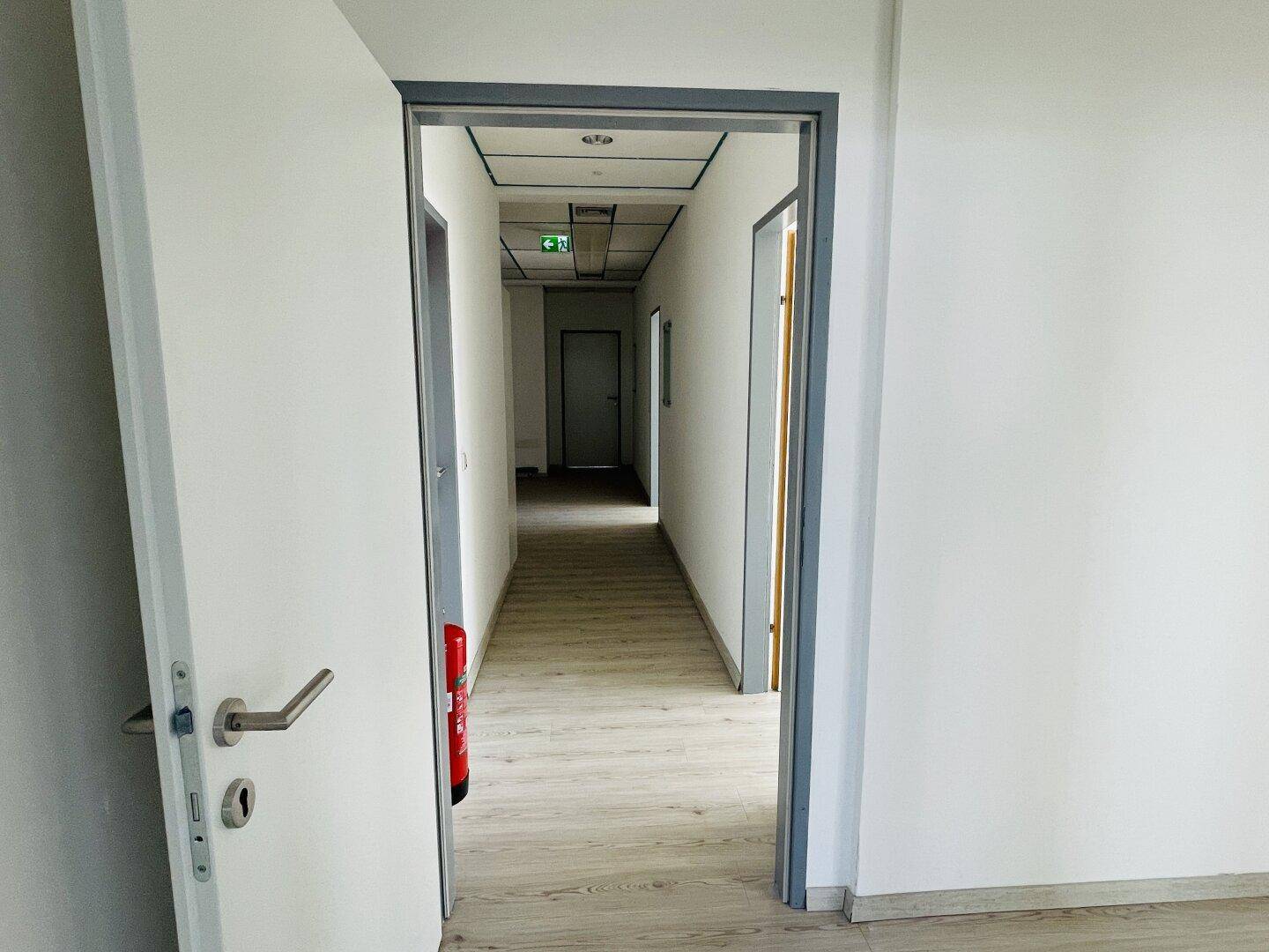 Langer, leerer Flur mit hellem Laminatboden und Rasterdecke, gesehen durch eine offene Tür.