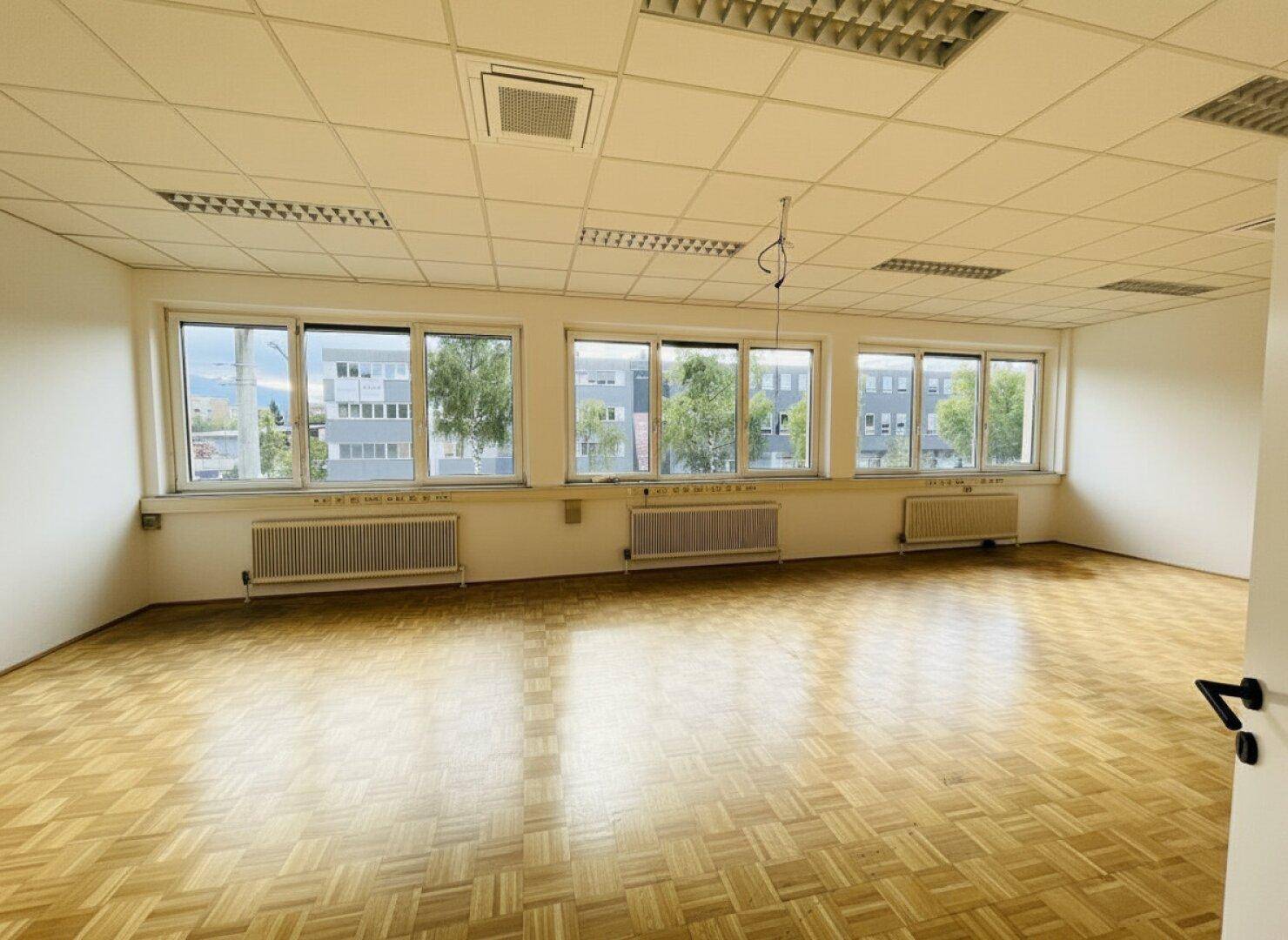 Geräumiges Büro mit Fischgrätparkett, Rasterdecke und großen Fenstern.