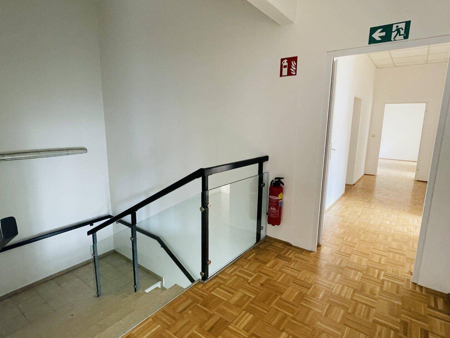 Treppenhausbereich mit Glasgeländer, Fischgrätparkett und Brandschutzvorrichtungen.