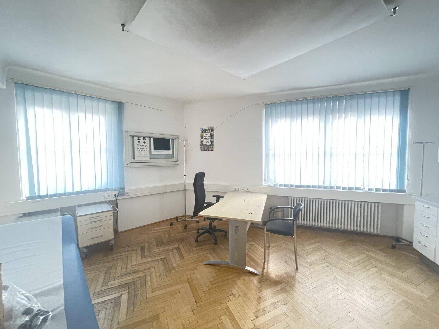 Helles Büro oder Behandlungszimmer mit zwei Fenstern, Heizkörpern und Fischgrätparkett.