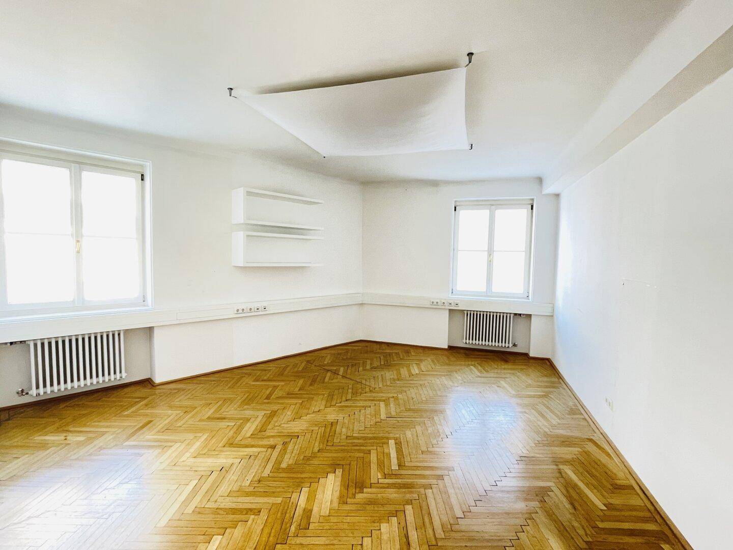 Leeres Büro mit Fischgrätparkett, weißen Wänden und zwei großen Fenstern.
