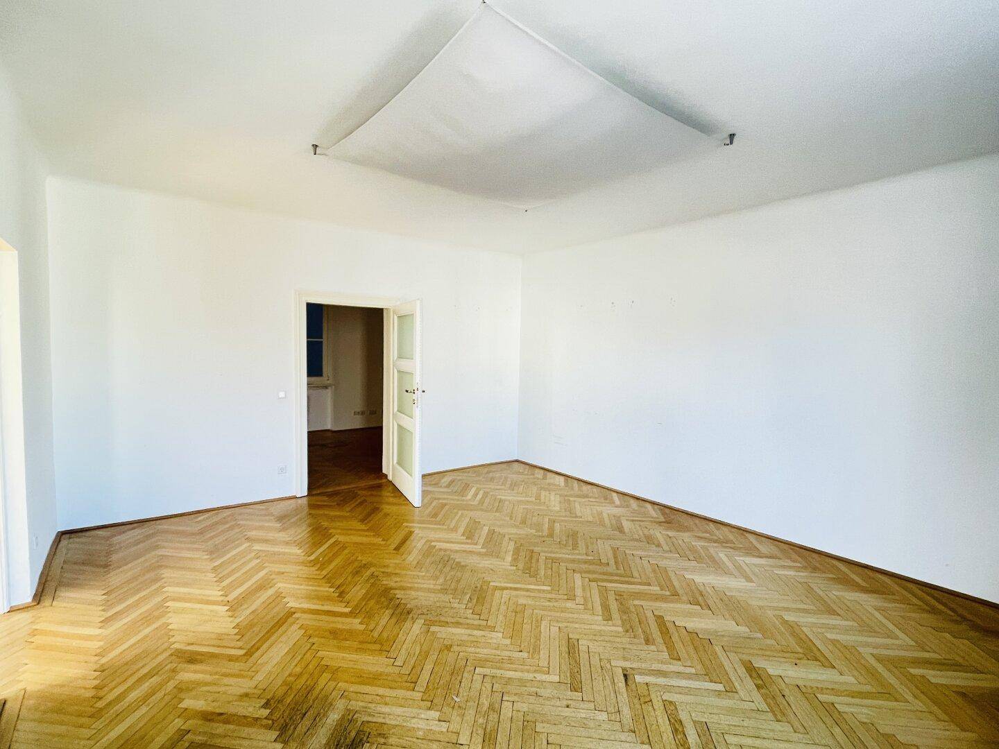 Geräumiges, leeres Zimmer mit Fischgrätparkett und weiß gestrichenen Wänden, bereit zur Nutzung.