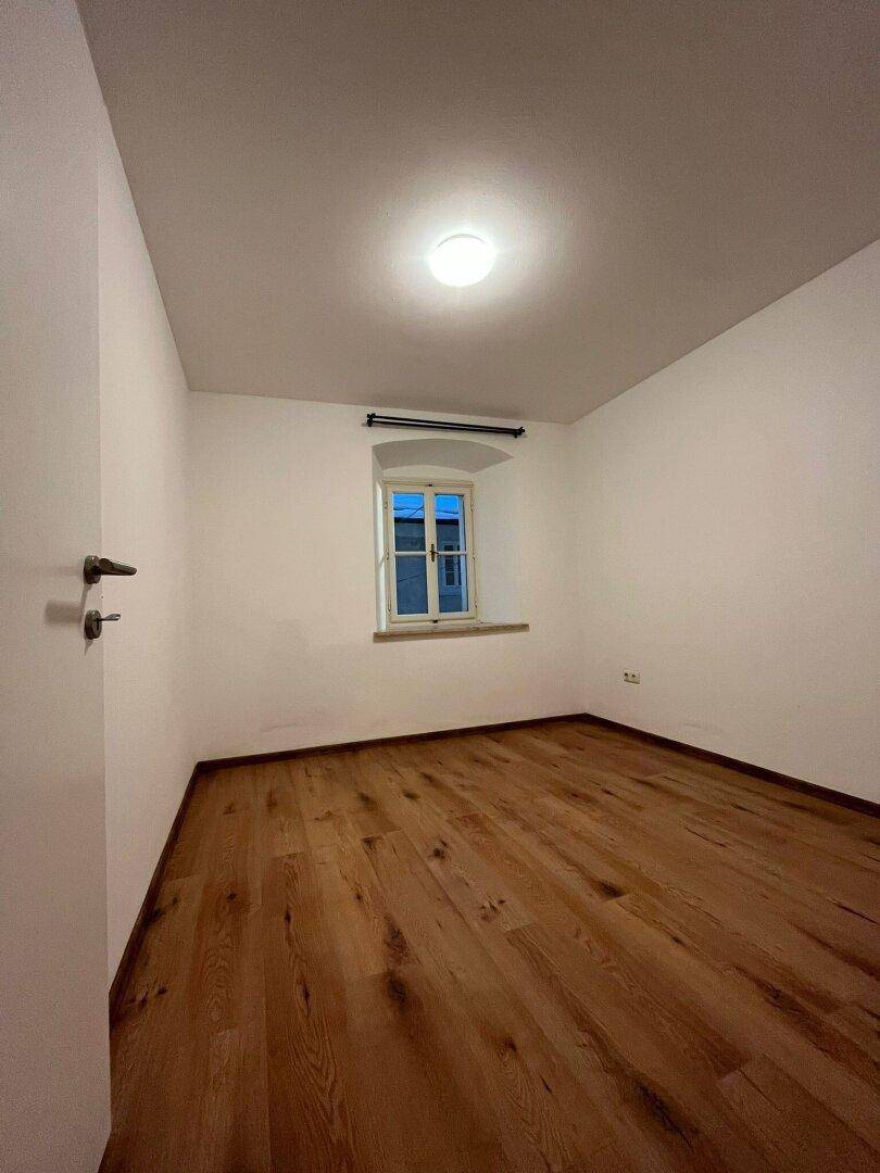Helles, leeres Zimmer mit Holzboden und einem kleinen Fenster, bereit zur individuellen Gestaltung.