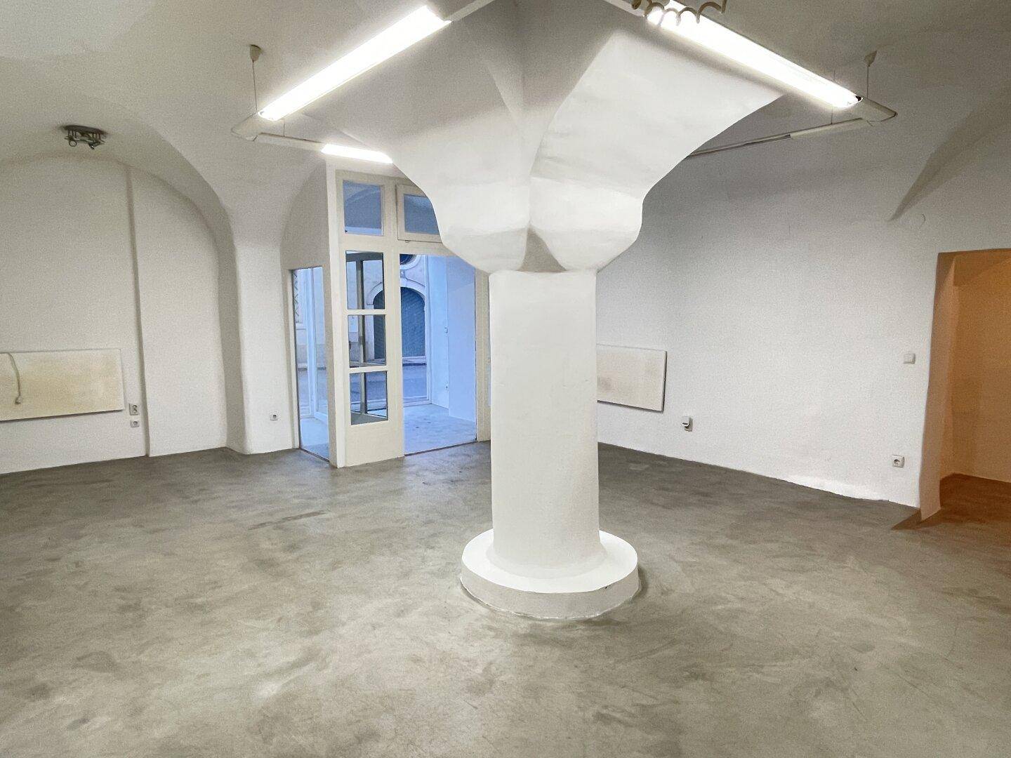Weitläufiger Raum mit markanter Säule, gewölbter Decke und großen Fenstertüren, die viel Licht hereinlassen.