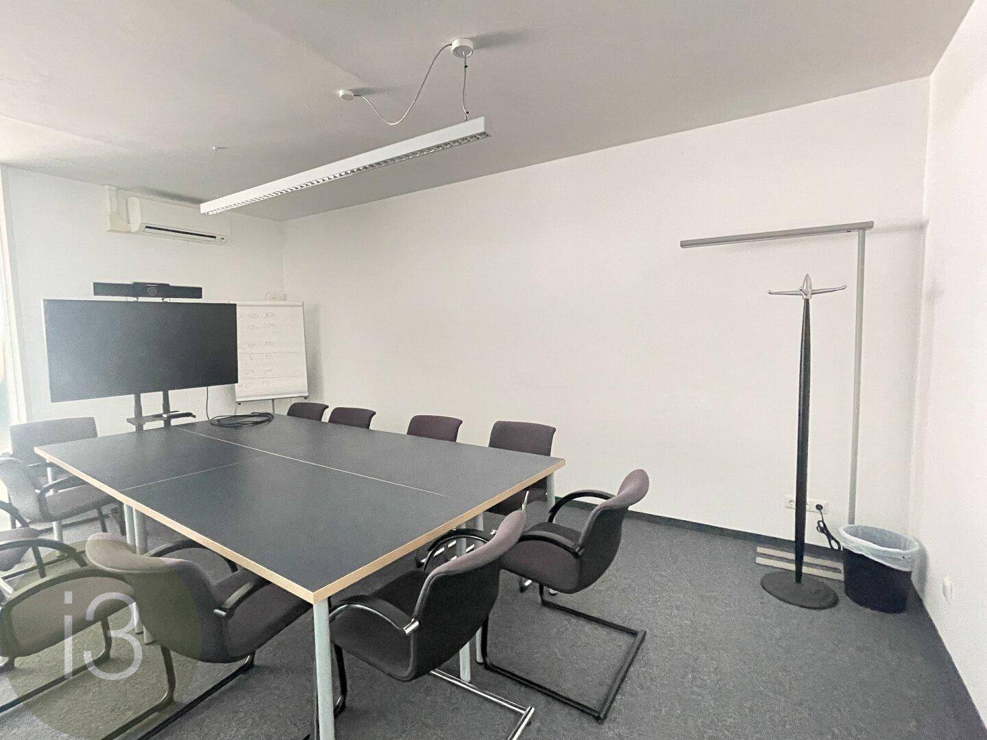 Konferenzraum mit großem Tisch, mehreren Stühlen, einem Wandbildschirm und Teppichboden für Besprechungen.