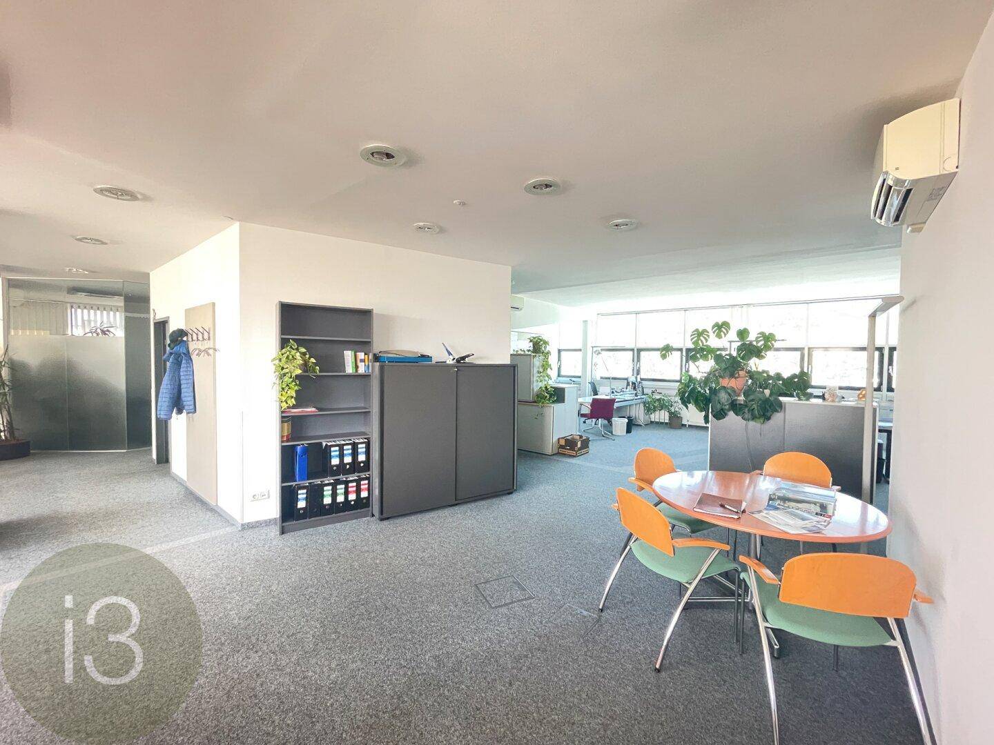 Offener Bürobereich mit Teppichboden, einer kleinen Sitzecke und Regalen, die den Raum strukturieren.